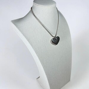 May include: A silver-toned necklace featuring a heart-shaped pendant with an intricate, dark-toned swirling filigree pattern. The pendant hangs from a smooth, round silver-toned chain that rests against the neck, showcasing a metallic finish.