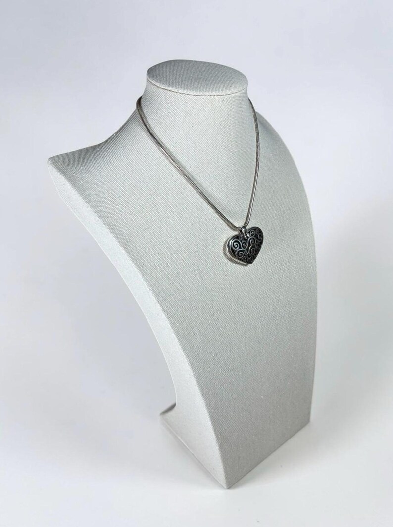 May include: A silver-toned necklace featuring a heart-shaped pendant with an intricate, dark-toned swirling filigree pattern. The pendant hangs from a smooth, round silver-toned chain that rests against the neck, showcasing a metallic finish.