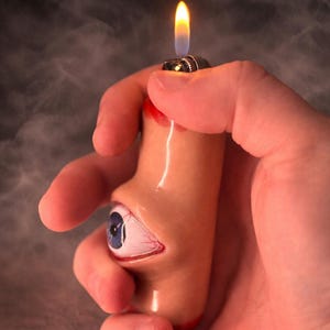 May include: A novelty lighter with a unique design. The lighter is shaped like a human eye and part of a face, with a blue iris and red veins. The flame is lit, and the lighter is held in a hand.