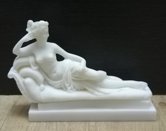 May include: White marble sculpture of a reclining woman on a base. The woman is wearing a flowing robe and has her arm resting on a pillow.