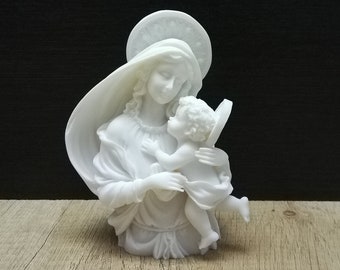 May include: White ceramic sculpture of the Virgin Mary holding a baby Jesus. The sculpture is on a light brown wooden surface.
