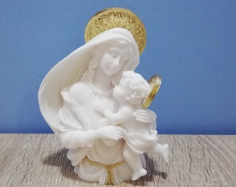 May include: A white statue of a woman holding a child, with a gold halo and accents. The sculpture is set against a blue background and rests on a wooden surface. The woman wears a draped veil.