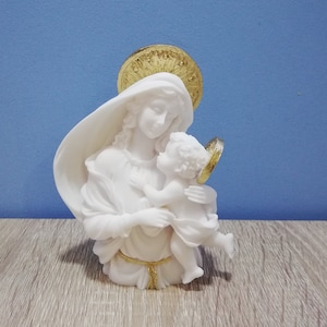 Può includere: Statua bianca di una donna che tiene in braccio un bambino, con un'aureola e dettagli dorati. La scultura è posta su uno sfondo blu e una superficie in legno. La donna indossa un velo drappeggiato.