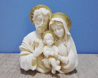 May include: A white ceramic figurine of the Holy Family.  The figures are depicted as a mother, father, and child. The mother and father are holding the child, who is wearing a simple robe. The figures are all wearing halos.