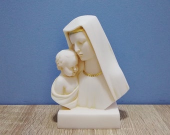 May include: A white bust sculpture of a woman wearing a veil, cradling a baby. The sculpture has a gold-colored necklace and trim on the veil. The sculpture is on a rectangular base, set against a blue background.