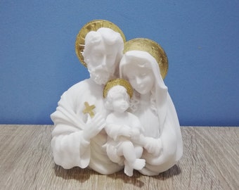 May include: A white marble sculpture of the Holy Family, featuring Joseph, Mary, and the infant Jesus. Each figure has a gold halo. Joseph holds a gold cross. The sculpture is set against a blue background and a wooden surface.