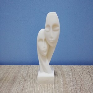 Op de afbeelding: Witte abstracte sculptuur van twee gezichten, een met gesloten ogen, de andere met open ogen, op een witte voet. De voet heeft de inscriptie "Devotion" erop.