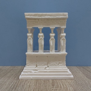 Puede incluir: Escultura de yeso blanco de un templo griego con cuatro figuras femeninas de pie en la entrada. El templo tiene una base detallada y un techo con una cornisa decorativa.