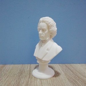 May include: A white bust sculpture of a man with curly hair and a stern expression. The sculpture is on a pedestal base and set against a blue background. The bust is a classical art piece.