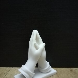 May include: A white marble sculpture depicting two hands joined in prayer. The hands, with detailed features, rest on a rectangular base. The sculpture is set against a dark backdrop, highlighting the white marble.