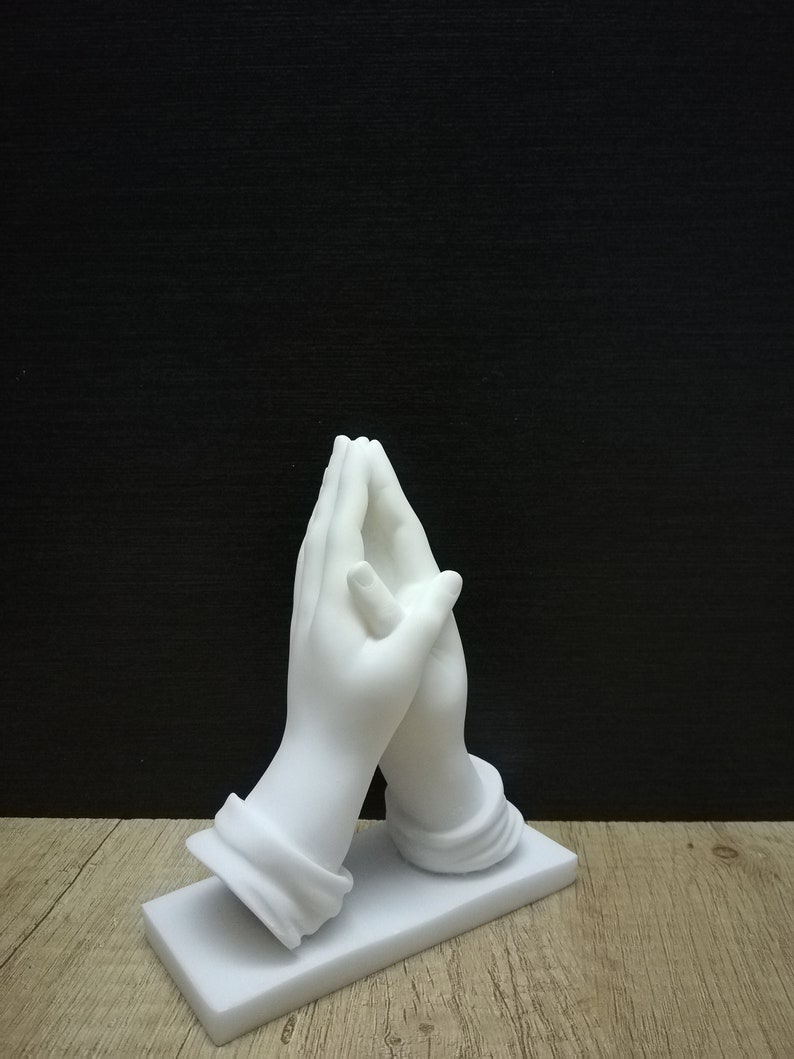 May include: A white marble sculpture depicting two hands joined in prayer. The hands, with detailed features, rest on a rectangular base. The sculpture is set against a dark backdrop, highlighting the white marble.