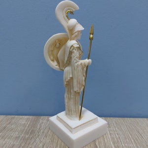 Może przedstawiać: Biała alabastrowa statuetka greckiej bogini wojowniczki z złotym hełmem, tarczą i wł&oacute;cznią. Statuetka stoi na białej podstawie.