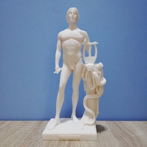 Könnte beinhalten: Eine weiße Statue einer nackten männlichen Figur, die eine Leier und einen kleinen Gegenstand hält. Die Statue steht auf einem rechteckigen Sockel mit der Inschrift "PHOEBUS". Der Hintergrund ist einfarbig hellblau.