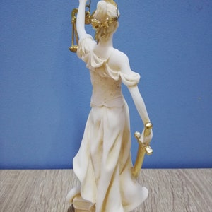 Puede incluir: Una estatua blanca de la figura de la Justicia, sosteniendo una espada y una balanza. La estatua est&aacute; hecha de un material blanco con detalles dorados. La estatua est&aacute; de pie sobre una superficie de madera.