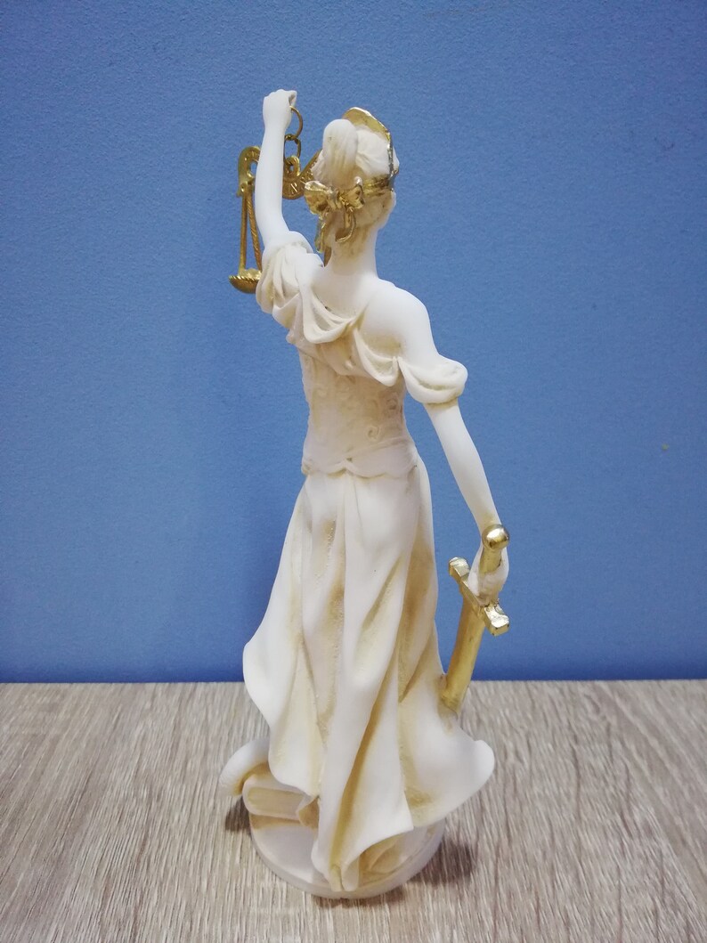Puede incluir: Una estatua blanca de la figura de la Justicia, sosteniendo una espada y una balanza. La estatua est&aacute; hecha de un material blanco con detalles dorados. La estatua est&aacute; de pie sobre una superficie de madera.