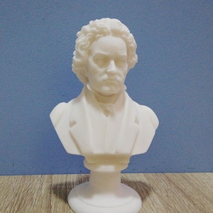 May include: A white bust of a man with curly hair and a serious expression. The sculpture is a classical style, with a detailed depiction of the subject's face and clothing. The bust is mounted on a round base.