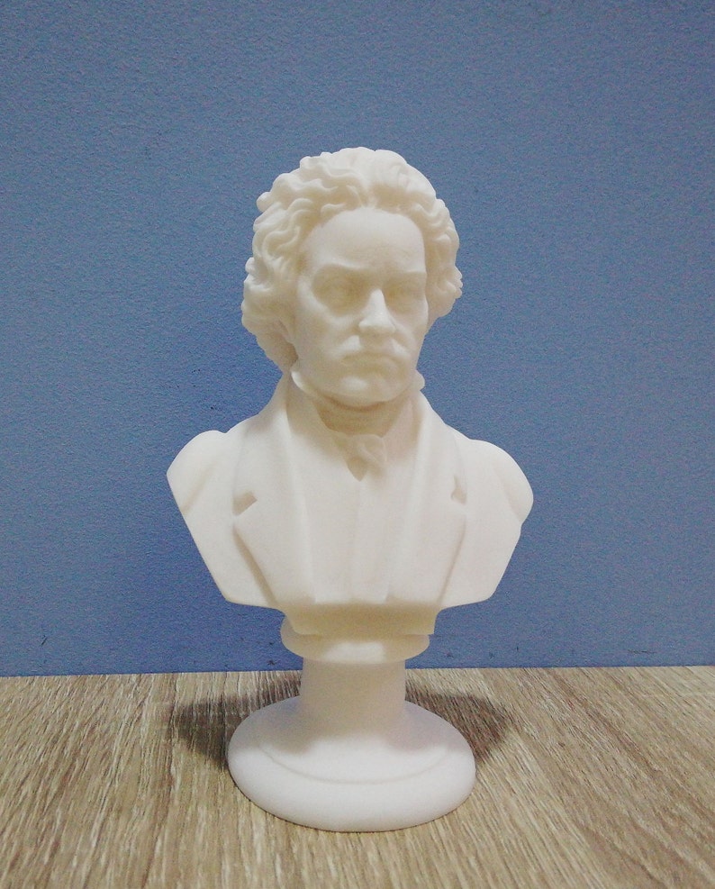 May include: A white bust of a man with curly hair and a serious expression. The sculpture is a classical style, with a detailed depiction of the subject's face and clothing. The bust is mounted on a round base.