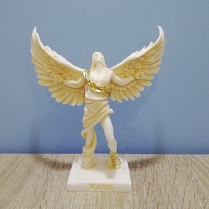 Peut inclure: Statue d'Icare en albâtre blanc avec des accents dorés, debout sur une base avec le mot "IKAROS" inscrit dessus.