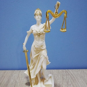 Puede incluir: Una estatua blanca de la figura de la Justicia, con los ojos vendados, sosteniendo una espada y una balanza. La balanza es dorada y la estatua est&aacute; sobre una base blanca.