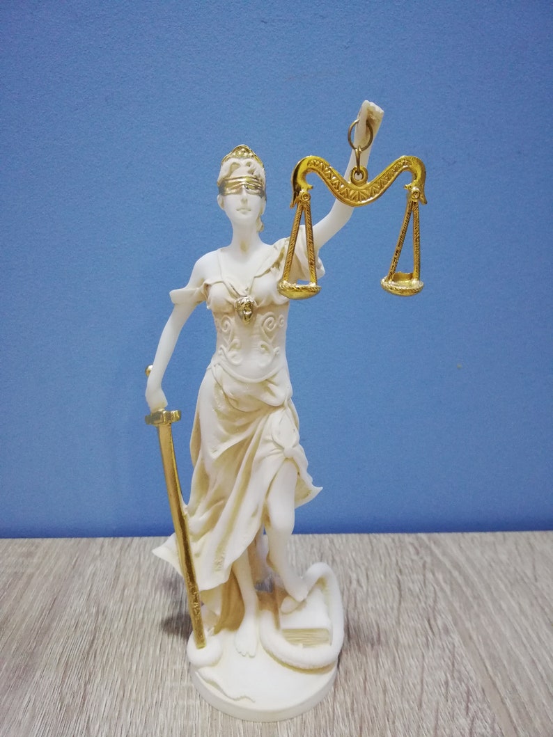 Puede incluir: Una estatua blanca de la figura de la Justicia, con los ojos vendados, sosteniendo una espada y una balanza. La balanza es dorada y la estatua est&aacute; sobre una base blanca.
