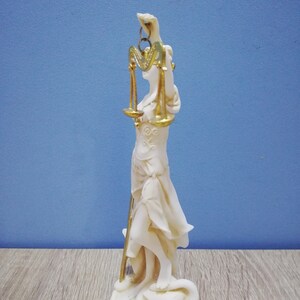 Puede incluir: Estatua blanca de la diosa griega de la justicia, Temis, sosteniendo una balanza en su mano derecha. La balanza es dorada y tiene un dise&ntilde;o en forma de coraz&oacute;n. La estatua est&aacute; de pie sobre un libro y una serpiente.