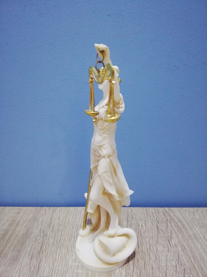 Puede incluir: Estatua blanca de la diosa griega de la justicia, Temis, sosteniendo una balanza en su mano derecha. La balanza es dorada y tiene un dise&ntilde;o en forma de coraz&oacute;n. La estatua est&aacute; de pie sobre un libro y una serpiente.