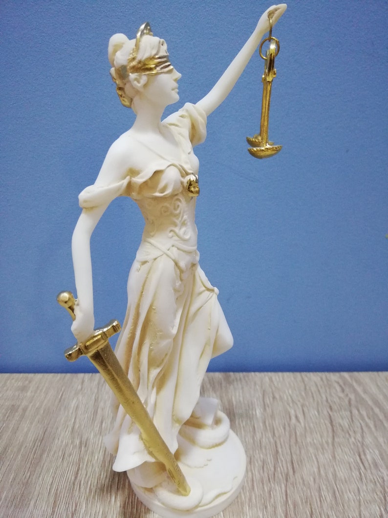 Puede incluir: Una estatua blanca de la diosa griega de la justicia, Temis, sosteniendo una espada y una balanza. La estatua est&aacute; vendada de los ojos y tiene una corona dorada.