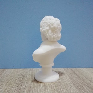 May include: A white bust sculpture of a man with curly hair, set against a blue background. The sculpture is on a pedestal base and appears to be made of a smooth, matte material. The bust is a classical art piece.