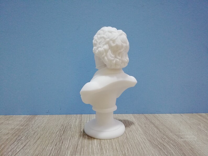 May include: A white bust sculpture of a man with curly hair, set against a blue background. The sculpture is on a pedestal base and appears to be made of a smooth, matte material. The bust is a classical art piece.