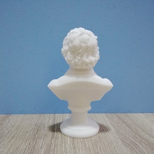 May include: A white bust sculpture with a detailed depiction of a person's head and shoulders. The sculpture has curly hair and is mounted on a pedestal base. The bust is set against a blue background.