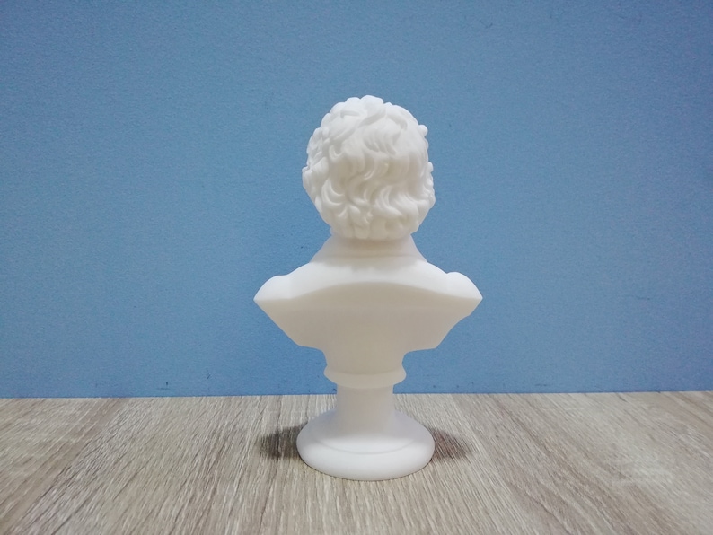 May include: A white bust sculpture with a detailed depiction of a person's head and shoulders. The sculpture has curly hair and is mounted on a pedestal base. The bust is set against a blue background.