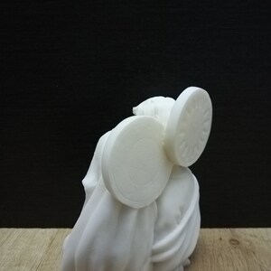 Puede incluir: Escultura de cer&aacute;mica blanca de una persona que lleva una capa con dos objetos circulares grandes en la cabeza.