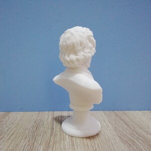 May include: A small, white bust sculpture of a person with curly hair and a classical style. The bust is on a round base and set against a blue background. The sculpture is made of a smooth, matte material.