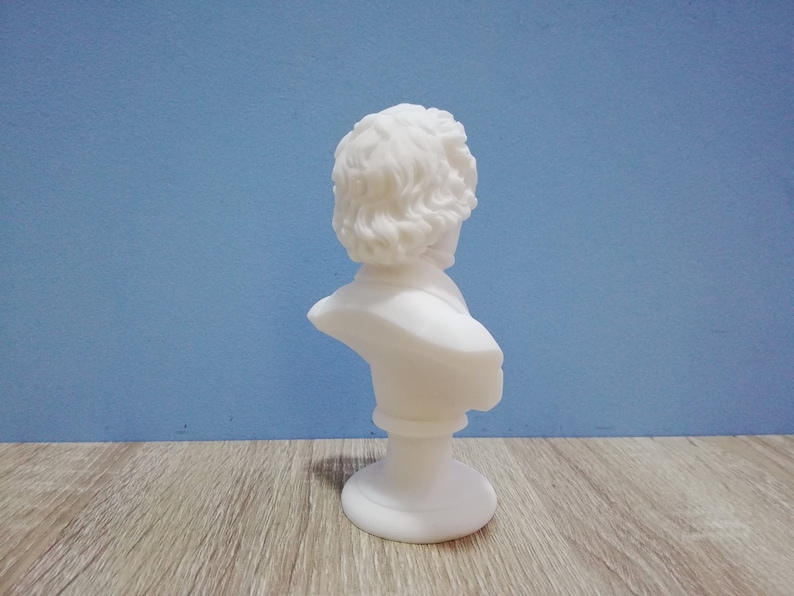 May include: A small, white bust sculpture of a person with curly hair and a classical style. The bust is on a round base and set against a blue background. The sculpture is made of a smooth, matte material.