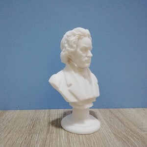 May include: A white bust of a man with curly hair and a jacket. The sculpture is on a round base and set against a blue background. The bust is a classical art piece.