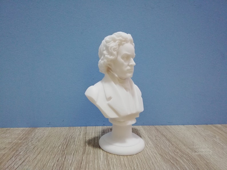 May include: A white bust of a man with curly hair and a jacket. The sculpture is on a round base and set against a blue background. The bust is a classical art piece.