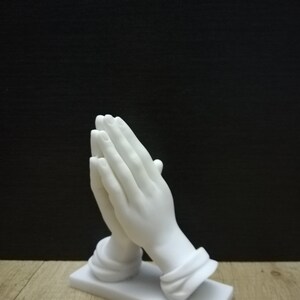 May include: A white marble sculpture of hands clasped in prayer. The hands are finely detailed, resting on a rectangular base. The background is a dark colour. The sculpture is a religious item.