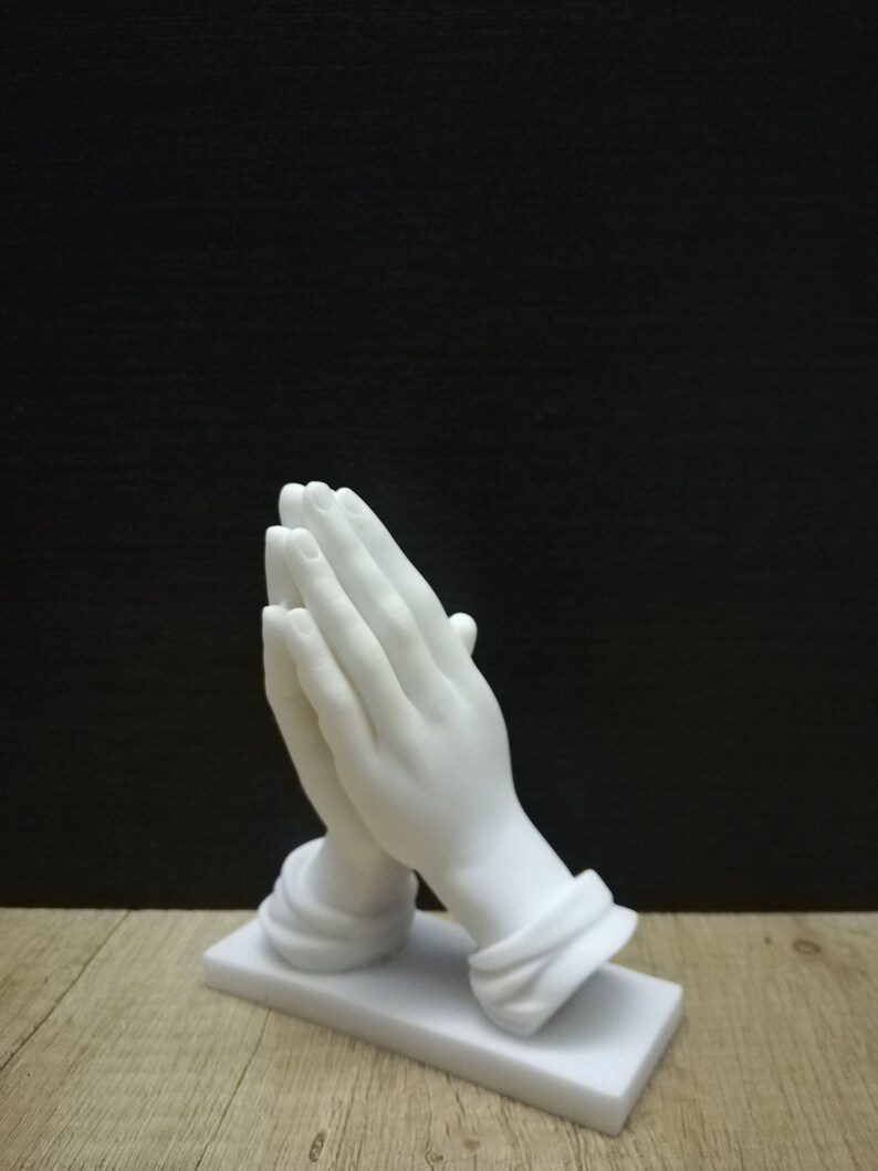 May include: A white marble sculpture of hands clasped in prayer. The hands are finely detailed, resting on a rectangular base. The background is a dark colour. The sculpture is a religious item.