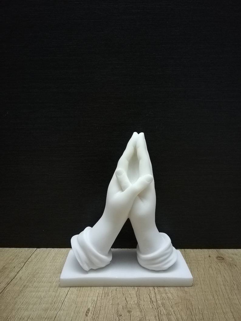 May include: A white sculpture depicting two hands in prayer, with fingers interlocked. The hands are set on a rectangular base. The background is a plain black, with a wooden surface below. The sculpture is a religious item.