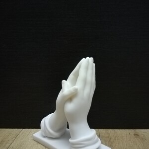 May include: A white sculpture of hands in prayer, set on a rectangular base. The hands are finely detailed. The background is black, and the sculpture is on a wooden surface.