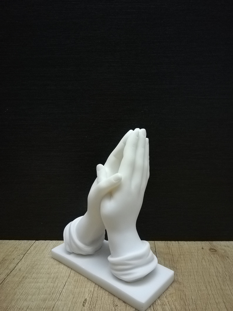 May include: A white sculpture of hands in prayer, set on a rectangular base. The hands are finely detailed. The background is black, and the sculpture is on a wooden surface.