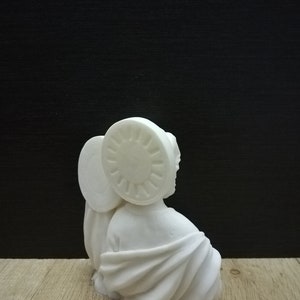 Puede incluir: Escultura de cer&aacute;mica blanca de una persona con una t&uacute;nica y un halo. La escultura est&aacute; sentada sobre una superficie de madera.