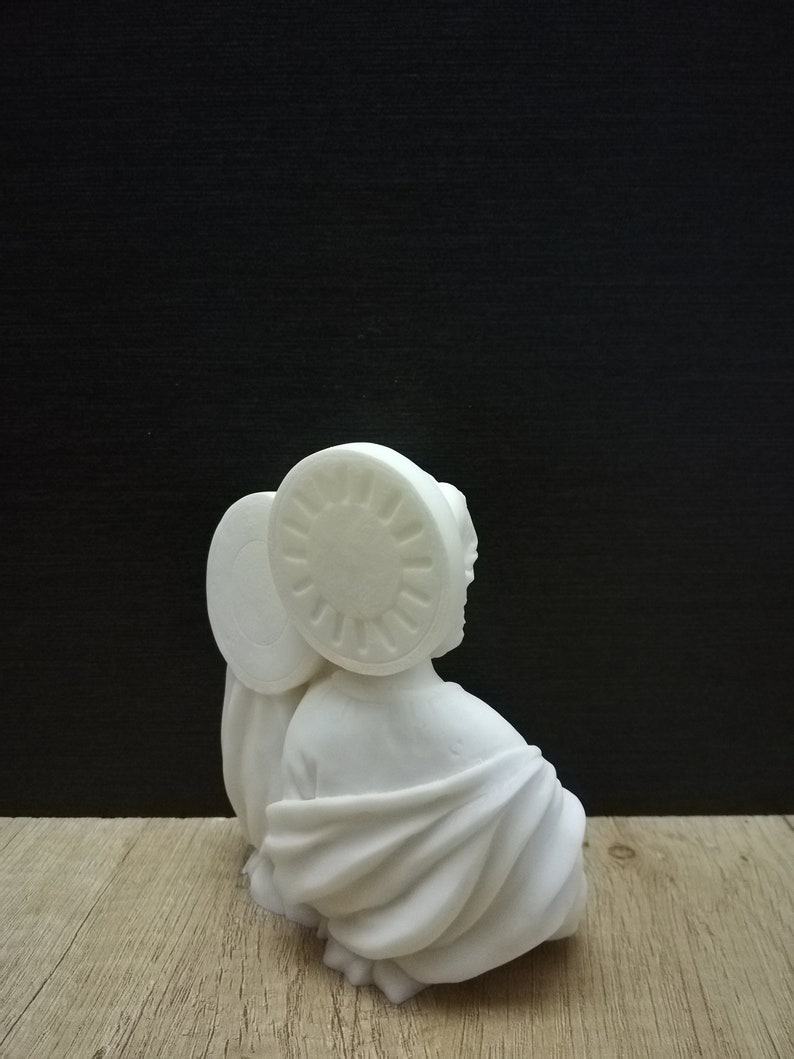 Puede incluir: Escultura de cer&aacute;mica blanca de una persona con una t&uacute;nica y un halo. La escultura est&aacute; sentada sobre una superficie de madera.