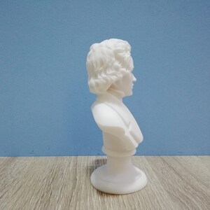 May include: A white marble bust of a person with curly hair and a high collar. The sculpture is mounted on a round base. The bust is set against a blue background and a wooden surface.