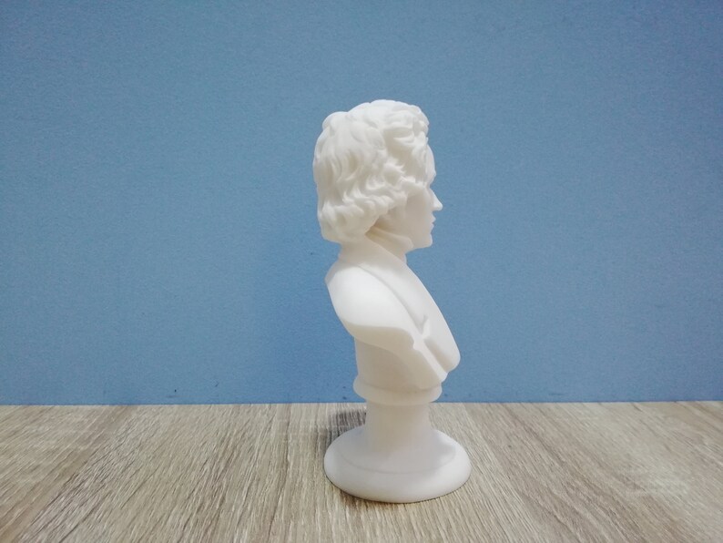 May include: A white marble bust of a person with curly hair and a high collar. The sculpture is mounted on a round base. The bust is set against a blue background and a wooden surface.