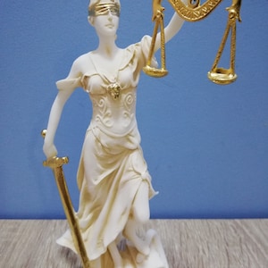 Puede incluir: Una estatua blanca de la diosa griega de la justicia, Temis, sosteniendo una espada y una balanza. La estatua est&aacute; vendada de los ojos y tiene una corona dorada.