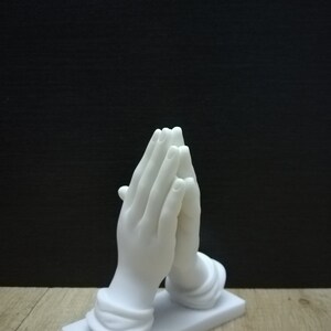 May include: A white sculpture depicting hands in prayer, with detailed fingers and a smooth finish. The hands are mounted on a rectangular base. The background is black, and the sculpture sits on a wooden surface.