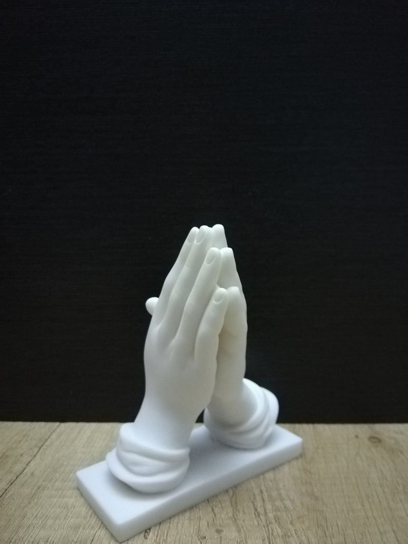 May include: A white sculpture depicting hands in prayer, with detailed fingers and a smooth finish. The hands are mounted on a rectangular base. The background is black, and the sculpture sits on a wooden surface.