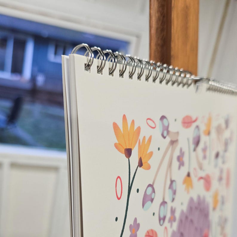 Up close of the binding for a 2026 wall calendar featuring colourful folk-art animal illustrations displayed in a flat lay. Wire-bound with premium thick paper, this illustrated monthly calendar showcases vibrant artwork for each month. Ideal for shoppers looking for a 2026 calendar, animal art calendar, home décor calendar, or unique artist-designed wall calendar.