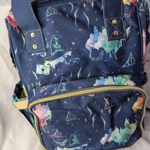 Può includere: Zaino blu navy con un motivo ripetuto di immagini a tema mago in stile acquerello. Lo zaino presenta una tasca frontale, cerniere dorate e spallacci e manici blu navy. Il design include un cappello da mago, occhiali e altri simboli magici.